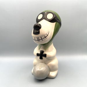 A ceramic figurine of a dog wearing a green aviator hat and goggles. The dog is sitting on a gray plane and has a white body with black eyes and a black mouth.
