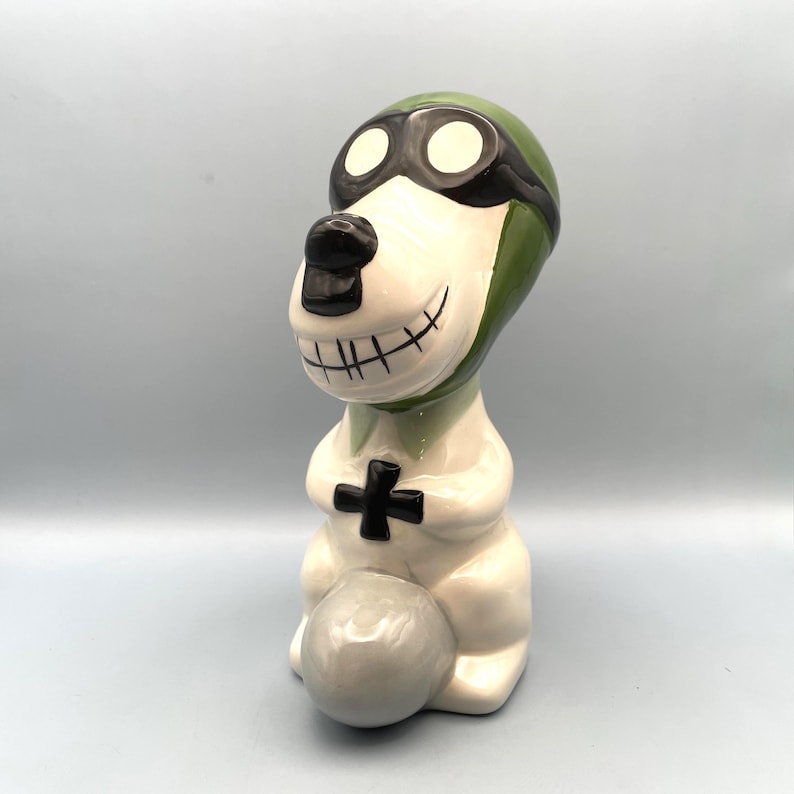 A ceramic figurine of a dog wearing a green aviator hat and goggles. The dog is sitting on a gray plane and has a white body with black eyes and a black mouth.