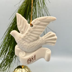 May include: A white ceramic dove ornament with outstretched wings, featuring a banner at the bottom with the year 1981 printed in brown text. The ornament hangs from a gold string against a background of green pine needles.