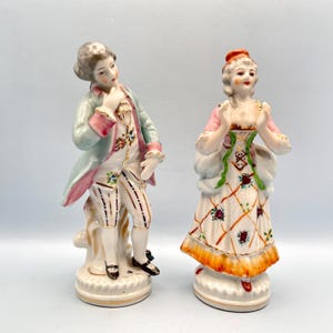 May include: Two porcelain figurines depicting a man and woman in 18th-century attire. The man wears a light blue coat with pink trim and striped pants. The woman is in a white dress with a floral pattern and orange accents.
