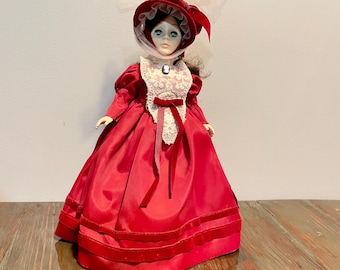 May include: A vintage porcelain doll in a long, red satin dress with velvet trim. The doll has a matching red bonnet with a white lace veil. The doll's dress has a white lace collar and a red ribbon at the waist. The doll has blue eyes and is a collectible item.