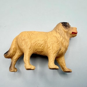 May include: A molded plastic dog figurine in a standing position, featuring a light tan coat with textured fur details, dark brown shading on the ears and tail, and a bright red painted mouth, captured from a side profile view.