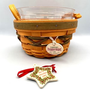 Może przedstawiać: Pleciony pomarańczowy kosz z przezroczystą szklaną wkładką i skórzanymi uchwytami. Kosz ma zielony pasek i etykietę z napisem "Longaberger Peppermint 1999 Tree Trimming". Obecna jest również gwiazdkowa ozdoba świąteczna z napisem "Merry Christmas 2001".