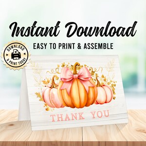 Peut inclure: Une carte de remerciement imprimable avec un fond en bois blanc. La carte représente trois citrouilles, dont une avec un nœud rose, et des accents en feuille d'or. Le texte "Merci" est imprimé en rose.