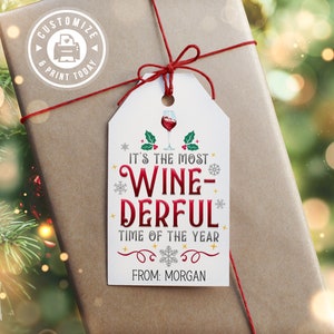 May include: White gift tag with red and gold text that reads "It's the most Wine-Derful time of the year."  A red wine glass with holly is above the text. The tag is tied with red string to a brown gift box.