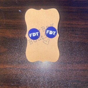 May include: A pair of blue stud earrings with the letters "FDT" in white. The earrings are on a brown, decorative tag with a heart and the text "Bay Boutique". The background is a dark, textured surface.