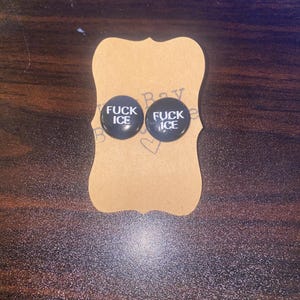 May include: A pair of black stud earrings with the text "FUCK ICE" in white lettering. The earrings are displayed on a brown, decorative card.
