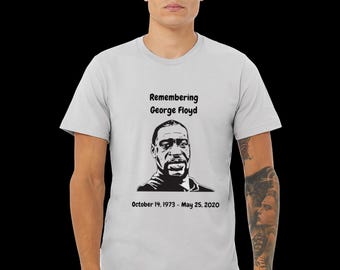 May include: A light gray t-shirt featuring a black and white portrait of George Floyd. The text "Remembering George Floyd" is above the portrait, with the dates "October 14, 1973 - May 25, 2020" below.