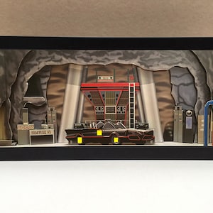 May include: A framed diorama of the Batcave from the 1960s Batman TV series. The diorama features a black Batmobile, a red and white Batcomputer, and a gray rock wall.