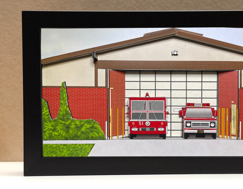 May include: A red fire truck with the number 51 on the side is parked in a fire station bay. The fire station is a paper model with a brown roof and red brick walls. There is a green bush in front of the fire station.