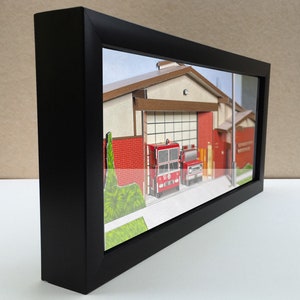 May include: A framed miniature model of a fire station with two red fire trucks parked in front. The fire station is made of paper and has a brown roof and red brick walls. The fire trucks are detailed and have the number 51 on the side.