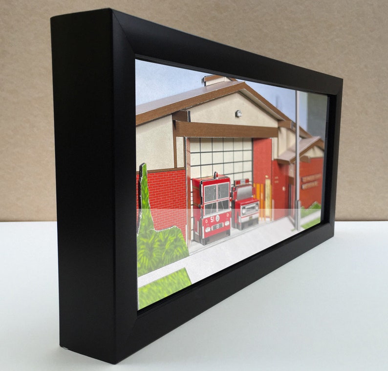 May include: A framed miniature model of a fire station with two red fire trucks parked in front. The fire station is made of paper and has a brown roof and red brick walls. The fire trucks are detailed and have the number 51 on the side.