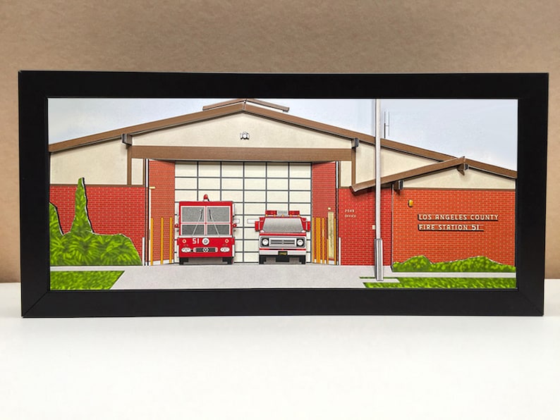 May include: A framed paper model of a Los Angeles County Fire Station 51, with a red fire truck and a red pickup truck parked in front of the building. The building is made of red brick with a brown roof and a white garage door. The model is set against a green lawn.