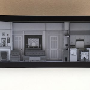 Può includere: Un diorama incorniciato nero che rappresenta un soggiorno, una cucina e un corridoio. Il diorama è fatto di carta e presenta un divano, un camino e elettrodomestici da cucina.