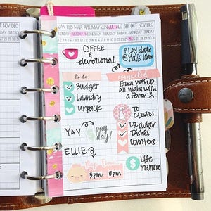 Peut inclure: Un agenda rose et blanc avec un horaire quotidien et une liste de choses à faire. L'agenda a des autocollants avec les mots "Coffee & Devotional", "Play Date", "To Clean", "Life Insurance", et "Nap Time".