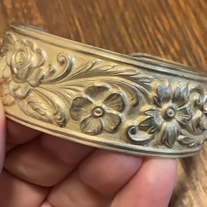 May include: A gold-toned cuff bracelet with intricate floral and leaf engravings. The detailed design features roses and other flowers, creating a vintage aesthetic. The bracelet is held in a hand.