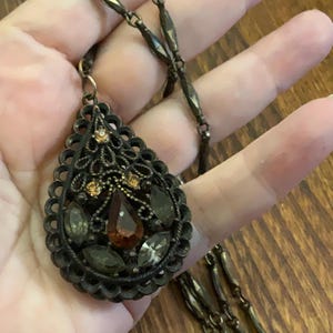 May include: An antique-style necklace with a teardrop pendant. The pendant features a filigree design with brown, amber, and clear gemstones. The chain is a dark bronze colour, and the necklace is displayed on a wooden surface.