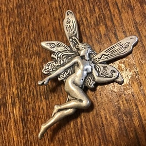 Pode incluir: Uma figura de fada prateada com as asas abertas. A fada é representada com cabelo solto e um design delicado e detalhado.