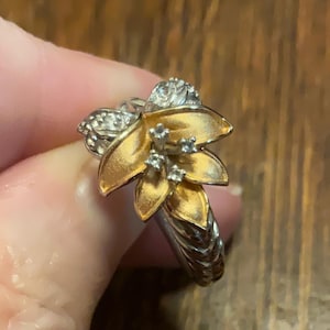 Puede incluir: Un anillo de oro y plata con un diseño de flor. La flor tiene cinco pétalos y está engastada con pequeños diamantes. La banda del anillo está trenzada.
