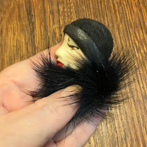 May include: A black and white porcelain brooch depicting a woman's face with red lips. The woman is wearing a black hat with a fur trim. The brooch is held in a hand.