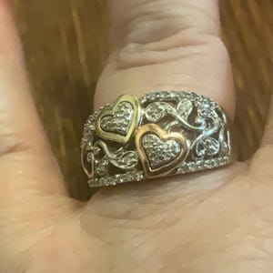 May include: A silver and gold ring with two hearts set with diamonds. The hearts are surrounded by a delicate floral design.