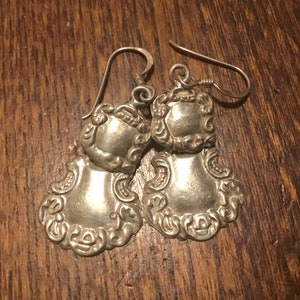 May include: A pair of silver earrings with a detailed, ornate design. The earrings are shaped like a teardrop with a scalloped edge and a floral pattern.