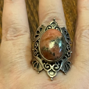 May include: A silver ring with an ornate design featuring a large oval-shaped pink and brown gemstone.