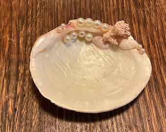 May include: A white seashell with a pink coral piece and pearls glued to the top. The shell is a decorative dish.