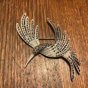 May include: A silver and black bird brooch with a long, pointed beak and intricate wings. The wings are made of rows of small, black stones.