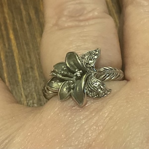 Könnte beinhalten: Ein silberner Ring mit einem Blumendesign. Die Blume hat eine silberne Mitte mit kleinen, klaren Steinen und die Blütenblätter sind silberfarben mit einer strukturierten Oberfläche.
