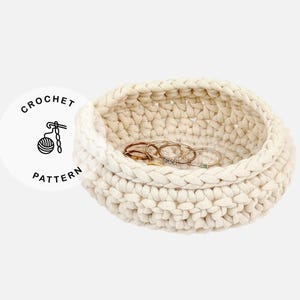 May include: A cream-colored, crocheted basket with a round shape, holding several rings. The basket is made of thick yarn and has a textured appearance. The image includes a circular graphic with the words "CROCHET PATTERN".