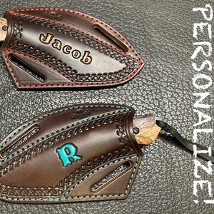 May include: Two brown leather knife sheaths with decorative stitching and personalized text. One sheath has the name "Jacob" in gold lettering, and the other has the letter "R" in turquoise. Both sheaths have a knife inside.