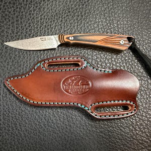May include: A small, rustic knife with a brown and black handle and a worn, silver blade rests above a brown leather sheath. The sheath has turquoise stitching and a stamped design. A black leather cord is attached to the knife.