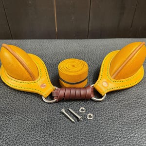 Può includere: Due maniglie in pelle gialla testurizzata con cuciture verdi, collegate da un anello in metallo rivestito in pelle marrone scuro. Include un rotolo di cinghia in pelle gialla abbinata, due viti in metallo argentato e due piccole rondelle argentate.
