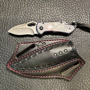 Może przedstawiać: Czarna skórzana pochwa na nóż z czerwonymi szwami i ozdobnym wzorem. Pochwa ma pętlę na pasek i zatrzask. Mały, srebrny scyzoryk z czarno-niebieskim wzorzystym uchwytem jest pokazany obok pochwy.