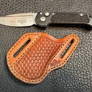 Può includere: Un coltello Microtech con lama argentata e manico nero, accanto a un fodero in pelle marrone. Il coltello presenta la scritta "UNDERWATER DEMOLITION TEAM" e "MICROTECH USA" sulla lama. Il fodero ha un motivo intrecciato.