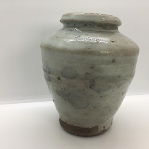 May include: A small, stout ceramic jar with a thick, off-white glaze that features subtle grey and brown speckles. The vessel has a wide, rounded body, a short neck, and a flared rim, with an unglazed, dark brown base showing the raw clay texture.