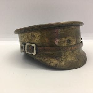 May include: A weathered brass military-style cap featuring a prominent visor and a thin, horizontal strap across the front. The metal surface displays a dark, aged patina with visible texture and a small, rectangular silver-toned buckle detail.
