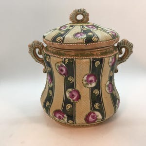 May include: A decorative ceramic jar with a lid, featuring a beige background with vertical dark green stripes. The jar is adorned with pink rose motifs and gold accents. It has two ornate handles and a circular handle on the lid.