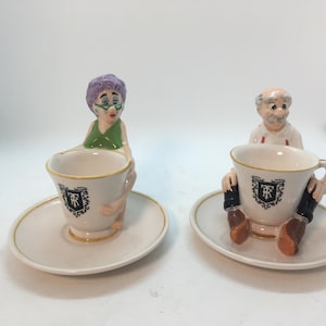 May include: A pair of whimsical ceramic teacups with saucers. One cup features a woman figure, the other a man. Both cups are white with gold trim and a black crest design. The saucers are also white with gold trim.