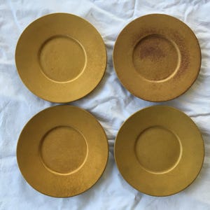 May include: Four gold-colored plates with a metallic finish are arranged on a white, textured surface. The plates have a circular shape with a slightly raised rim and a central indentation. One plate shows signs of wear.