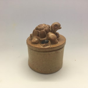 May include: A small, round, tan ceramic jar with a lid, featuring a turtle figurine. The turtle, with a textured shell, sits atop the lid. The jar is a simple, yet charming, decorative piece, perfect for storing small items.