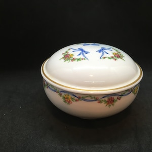 May include: A white porcelain trinket box with a gold rim and a floral design featuring blue bows and yellow, red, and pink flowers.