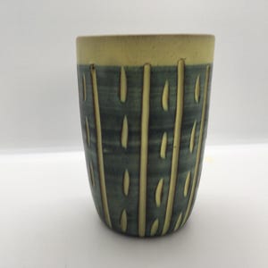 May include: A handmade ceramic tumbler with a cylindrical shape. The upper rim is a light yellow-green, while the body is a darker teal with vertical, textured, yellow-green lines. The tumbler is suitable for beverages.