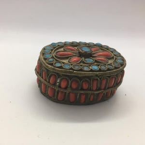 May include: An antique, oval jewellery box with a brass frame. The box is adorned with red and turquoise-coloured stones. The lid features a floral design with turquoise stones. The sides are decorated with red stones.