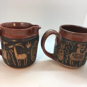 May include: Two ceramic mugs with brown glaze and black and brown designs. One mug has a design of an animal with antlers and the other mug has a design of a bug and a person riding an animal.