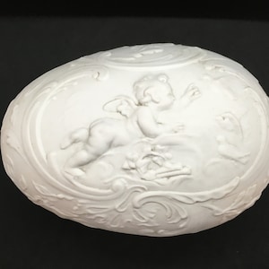 May include: White ceramic egg-shaped ornament with an embossed design of a cherub with wings and a floral pattern.