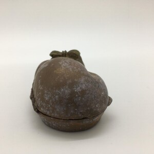 May include: A small, antique-style trinket box in the shape of a fruit, possibly a peach or apple. The box is a muted brown color with white speckling, and a bow detail on top. The box is closed, with a visible seam.