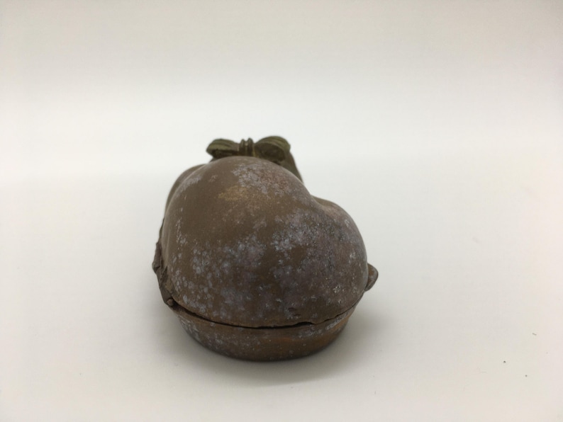 May include: A small, antique-style trinket box in the shape of a fruit, possibly a peach or apple. The box is a muted brown color with white speckling, and a bow detail on top. The box is closed, with a visible seam.