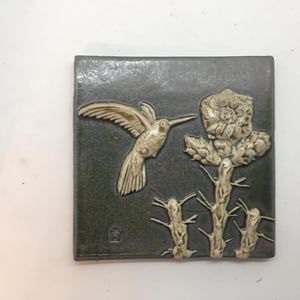 May include: A ceramic tile with a raised design of a hummingbird in flight, hovering near a cactus flower. The tile has a dark green background and the hummingbird and flower are a light cream color.